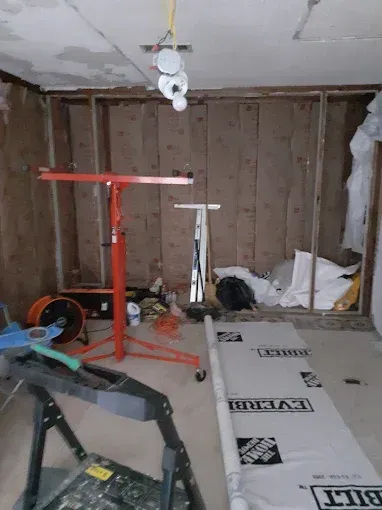 A room under renovation with exposed wall studs, insulation, a red drywall lift, and construction materials on the floor.