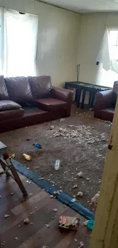 A living room with brown leather furniture and debris scattered across the carpeted floor.