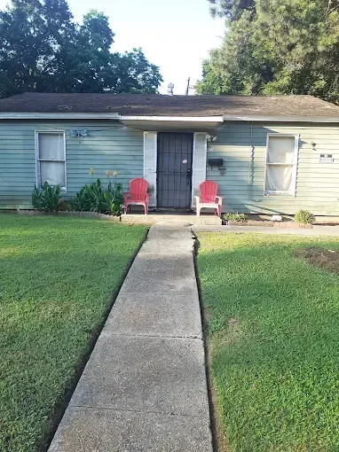 A pale green, single-story house with two red chairs on the porch and a concrete walkway leading to the front door.