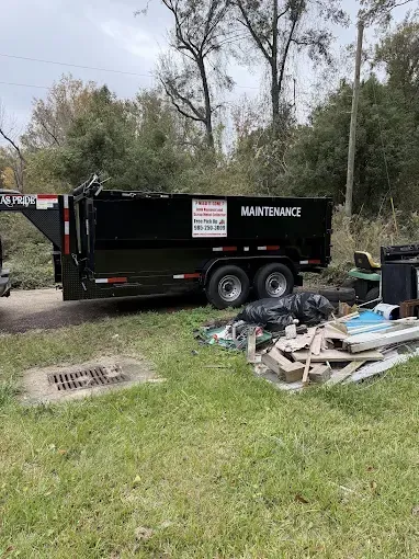 A black dump trailer parked on a grassy lot next to a pile of discarded wood, debris, and plastic bags.