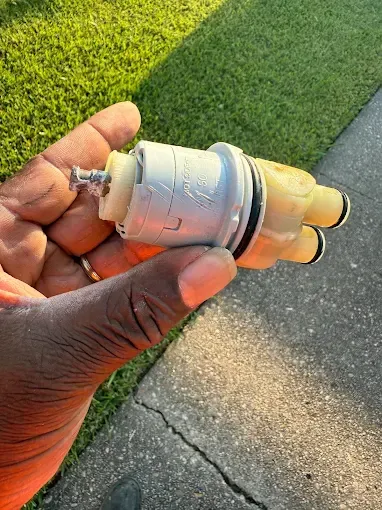 A person holds a white and yellow plastic plumbing faucet cartridge against an outdoor grassy background.