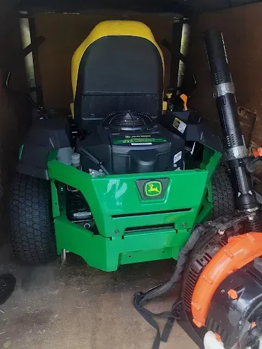 A green John Deere zero-turn lawn mower parked in a shed next to an orange leaf blower.