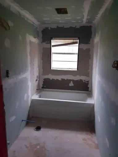 A bathroom under renovation with green drywall, a white bathtub, and a window centered above the tub.