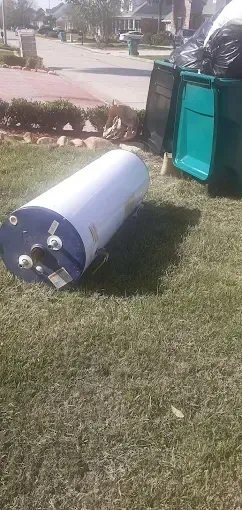 A white cylindrical water heater lies on the grass next to a green rolling trash bin outside a suburban home.