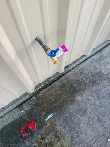 A brass-colored water spigot with a blue handle and a small pink tag mounted on a tan metal wall above concrete.