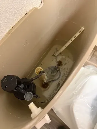 The interior of an open beige toilet tank, showing the flush valve, fill valve, and disconnected white plastic flush lever.
