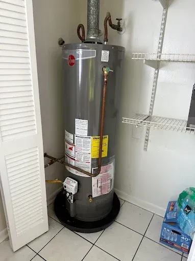 A gray residential water heater sits in a white closet on a black drip pan, with pipes and wire shelving visible.
