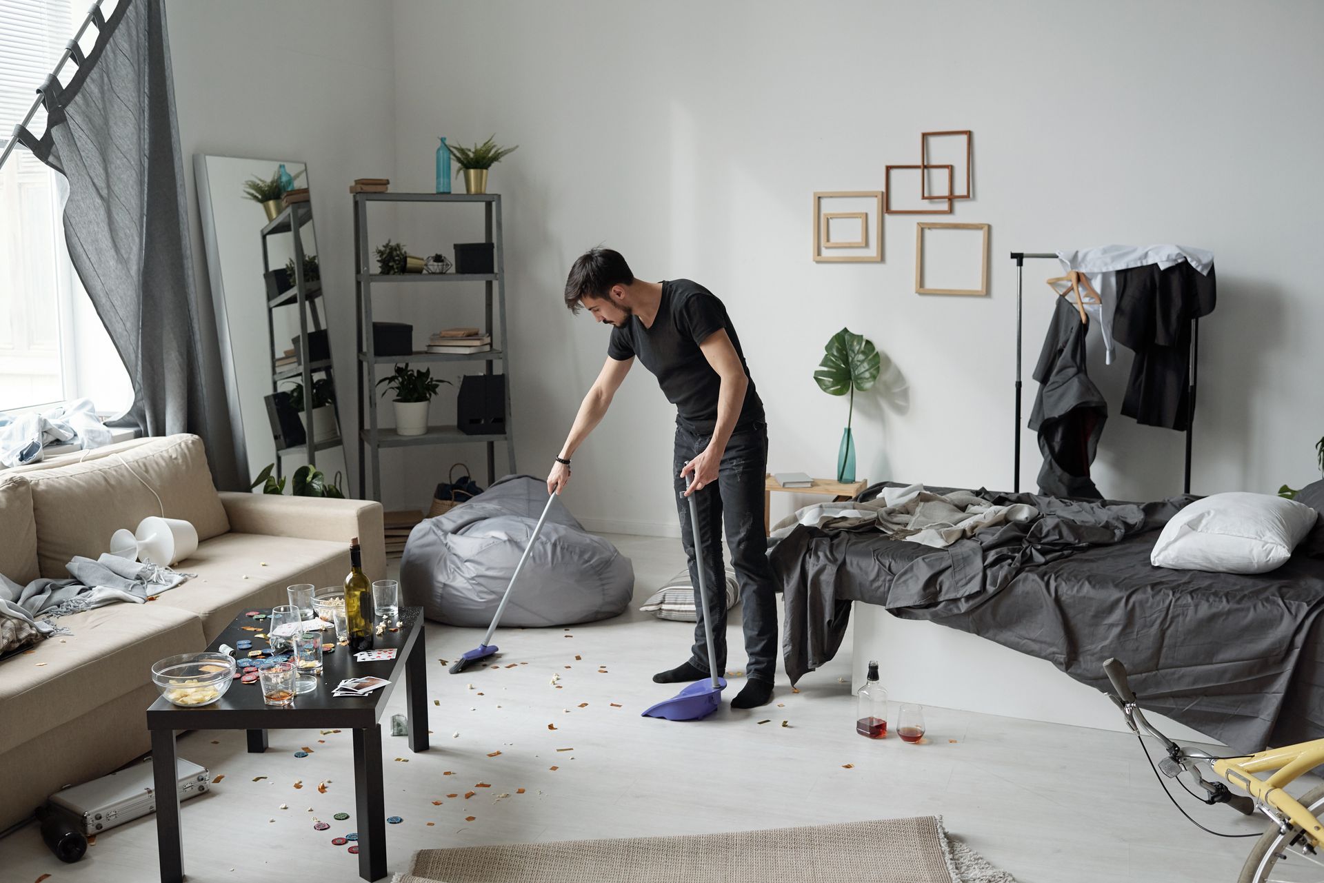 A person in a black shirt and pants sweeps debris off the floor of a messy room with a broom and dustpan.