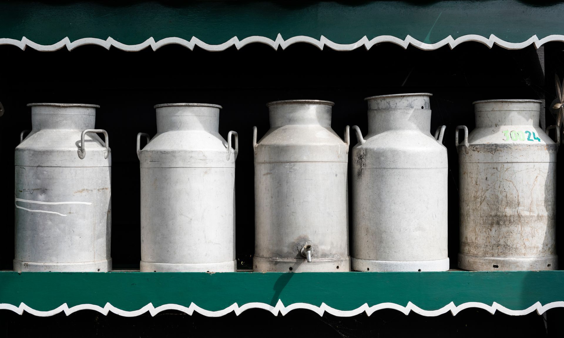Five silver milk cans lined up on a green shelf with a black background.