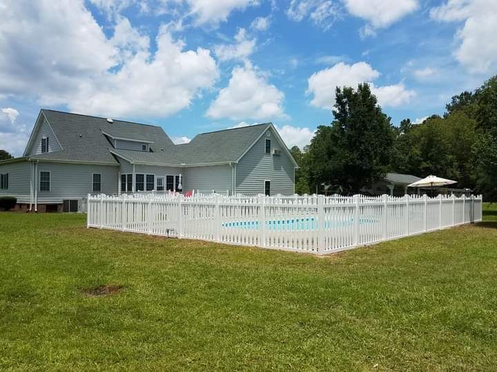 White fenced-in pool in a grassy backyard with a large light gray house and blue sky.