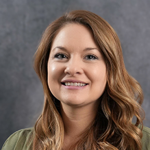 A woman with long brown hair is smiling and wearing a green shirt.