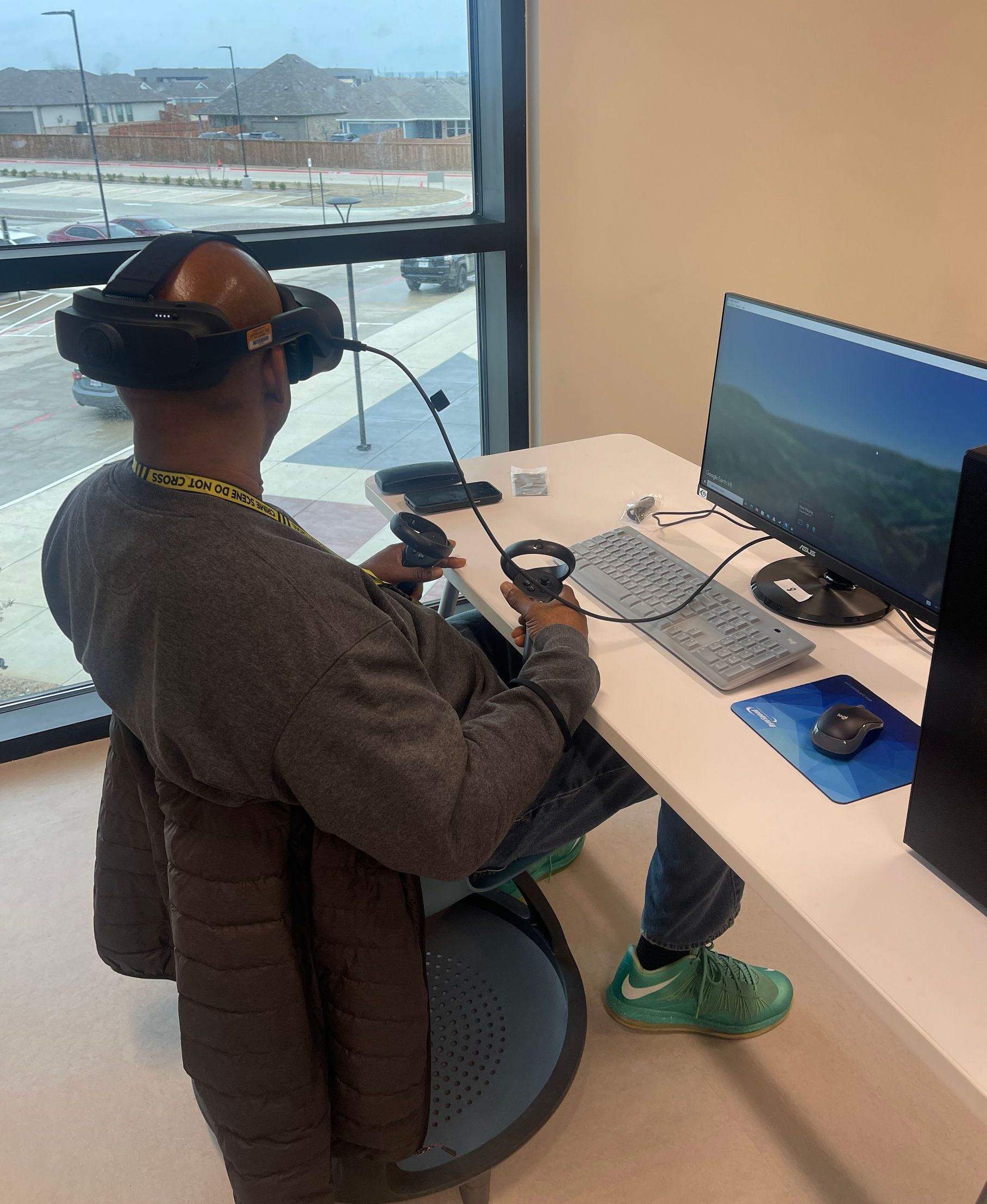 A man wearing a virtual reality headset is sitting at a desk with a computer.