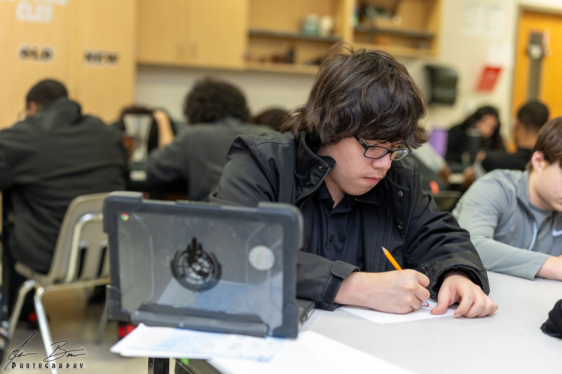 A boy is sitting at a desk in a classroom writing on a piece of paper.
