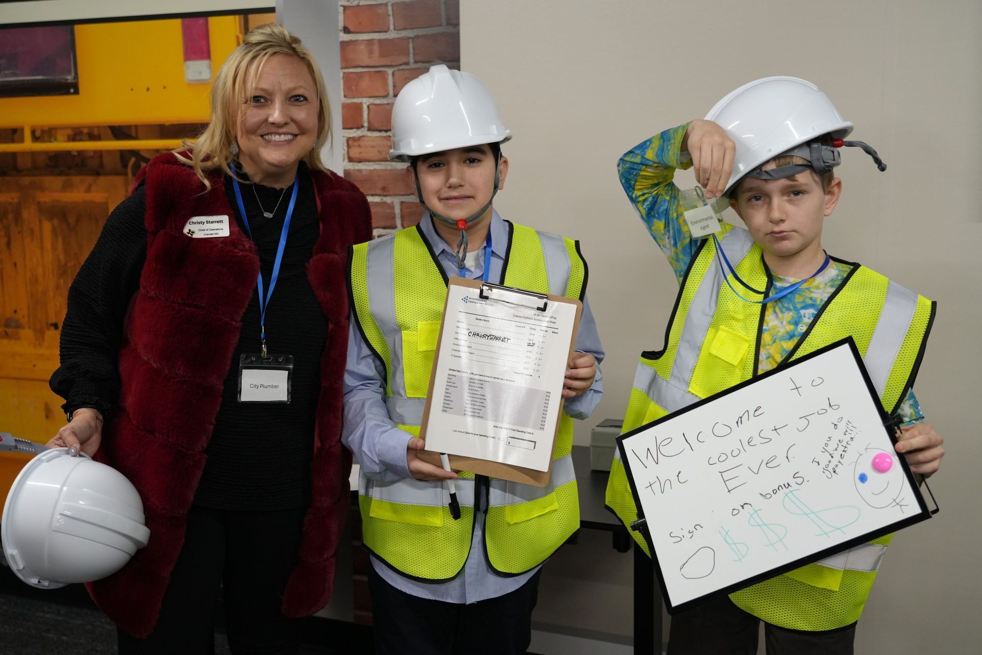 A woman is standing next to two children wearing hard hats and safety vests.