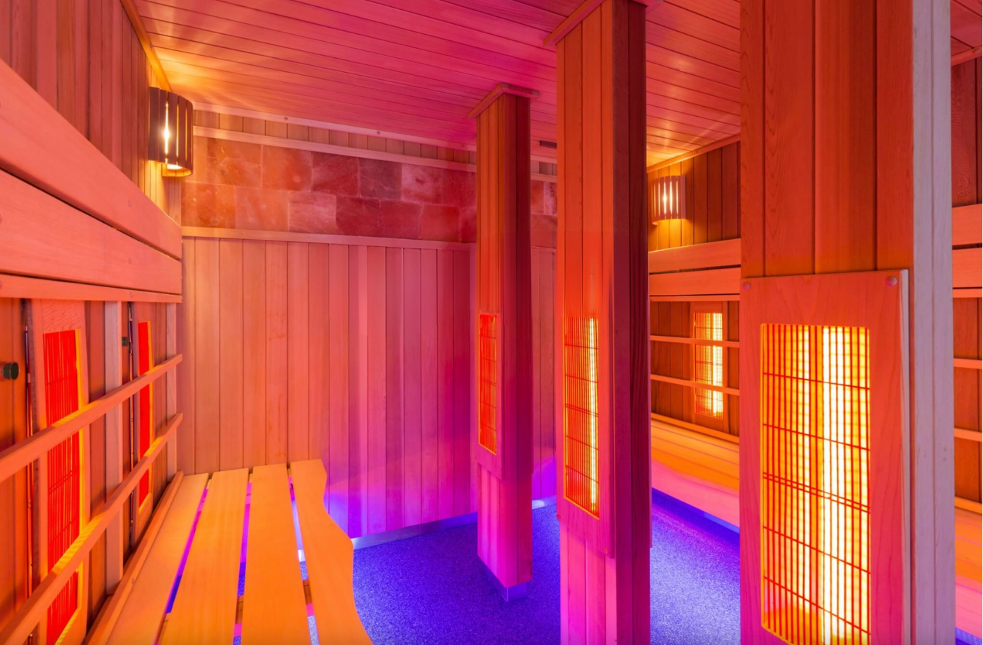 Intérieur d'un sauna avec bancs en bois, murs et panneaux chauffants infrarouges, éclairé par une lumière rouge et violette.