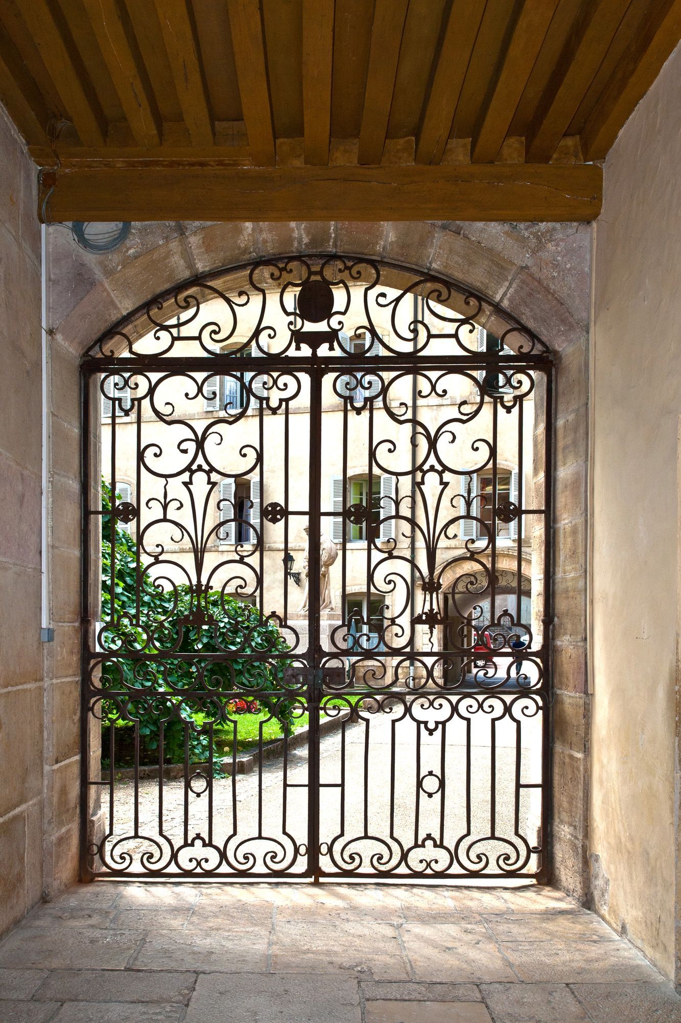 Cancello in ferro decorato che conduce a un cortile con edifici e vegetazione.