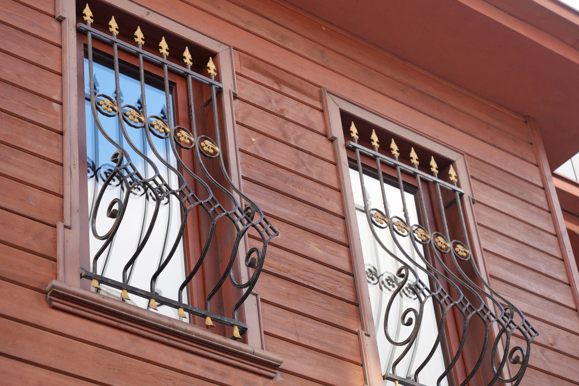 Due finestre con inferriate in metallo nero decorato e dettagli dorati su un edificio in legno marrone-rossastro.