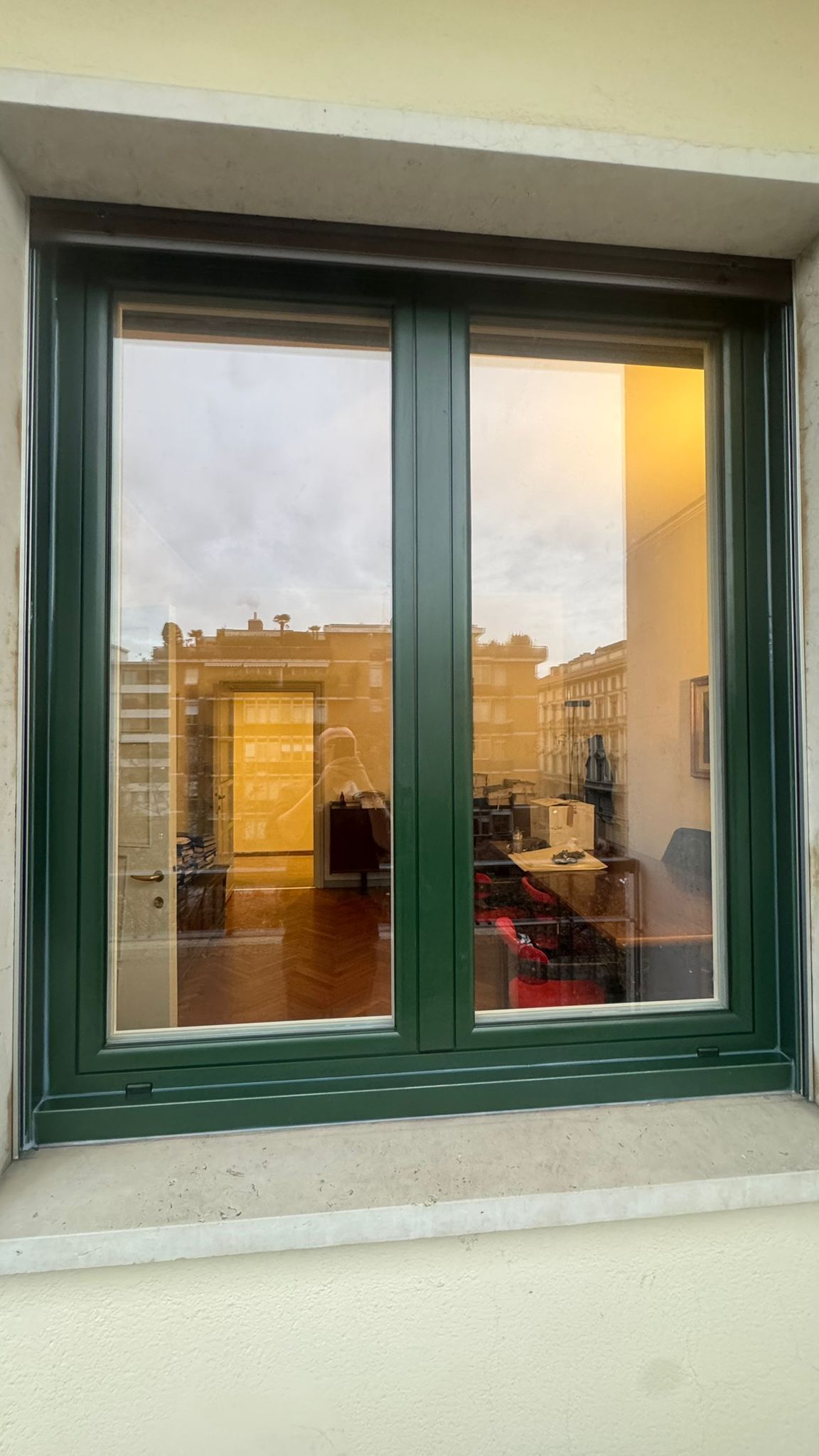 Finestra con cornice verde che riflette uno spazio interno e un paesaggio urbano esterno con edifici e luce solare.