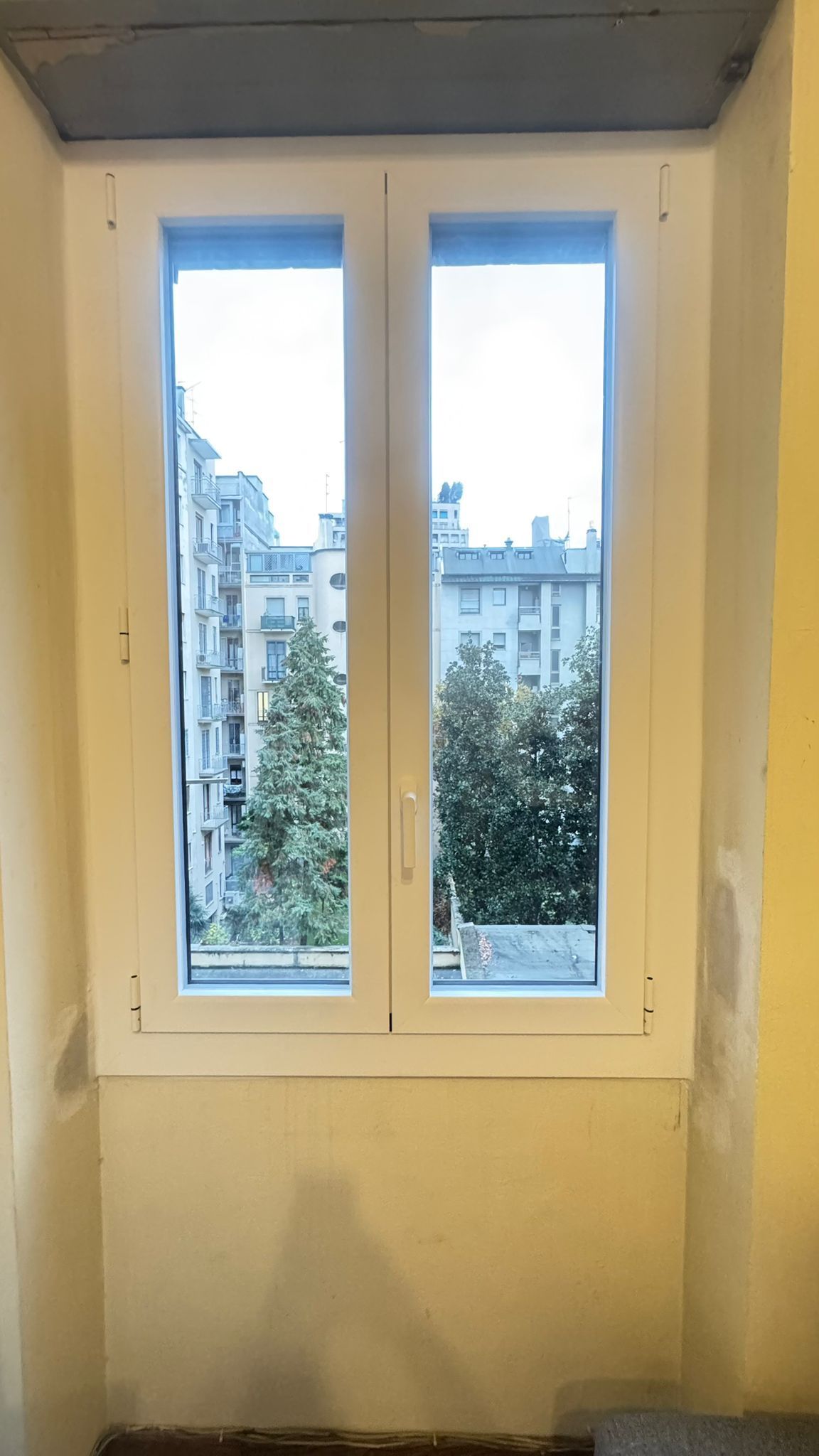 Doppia finestra con cornice bianca che si affaccia su edifici e alberi, incastonata in una parete giallo chiaro.