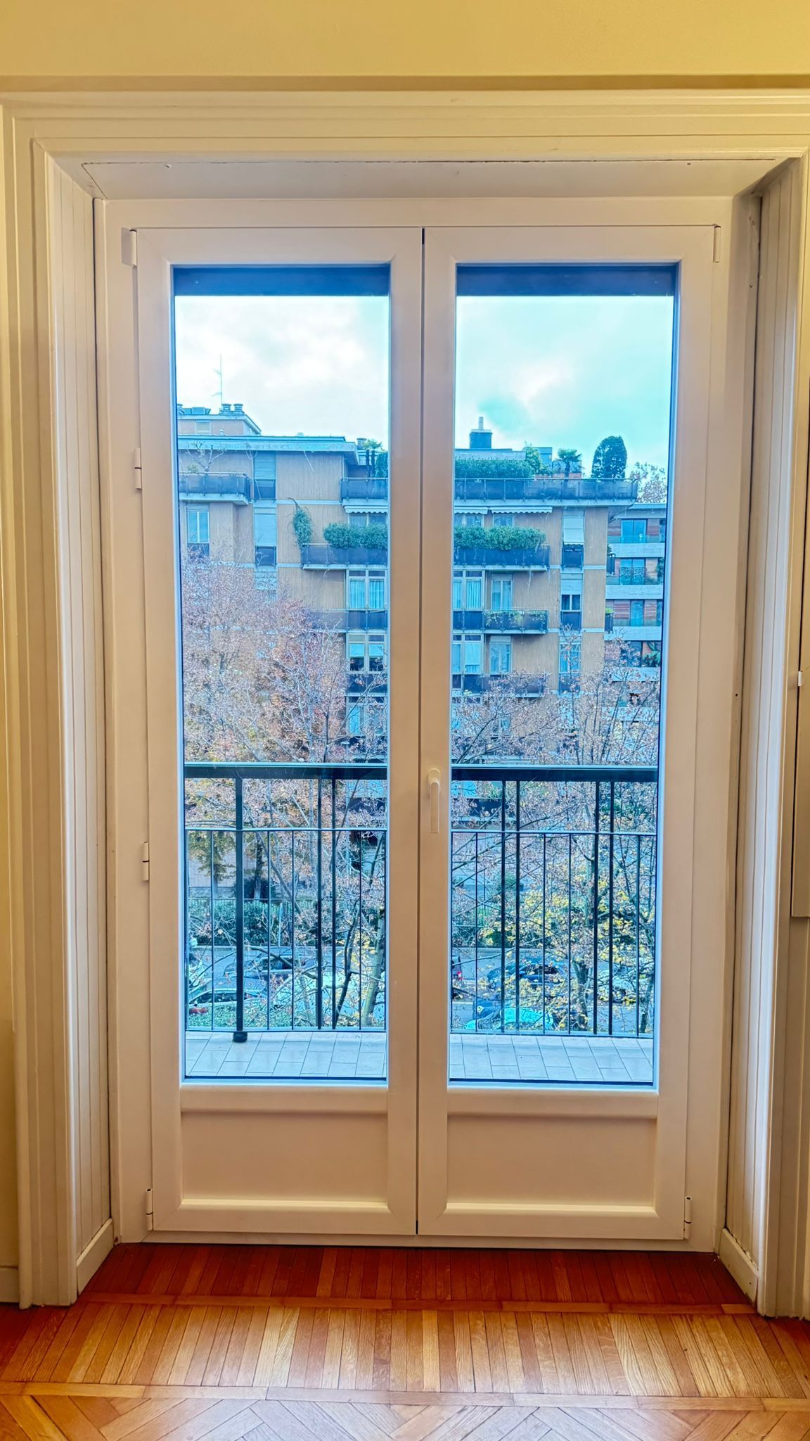 Le doppie porte bianche si aprono su un balcone con ringhiere nere e vista su condomini e alberi.