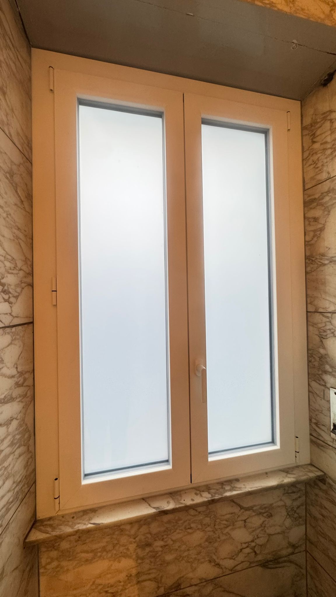 Finestra con cornice bianca e vetro smerigliato incastonata nella parete effetto marmo.
