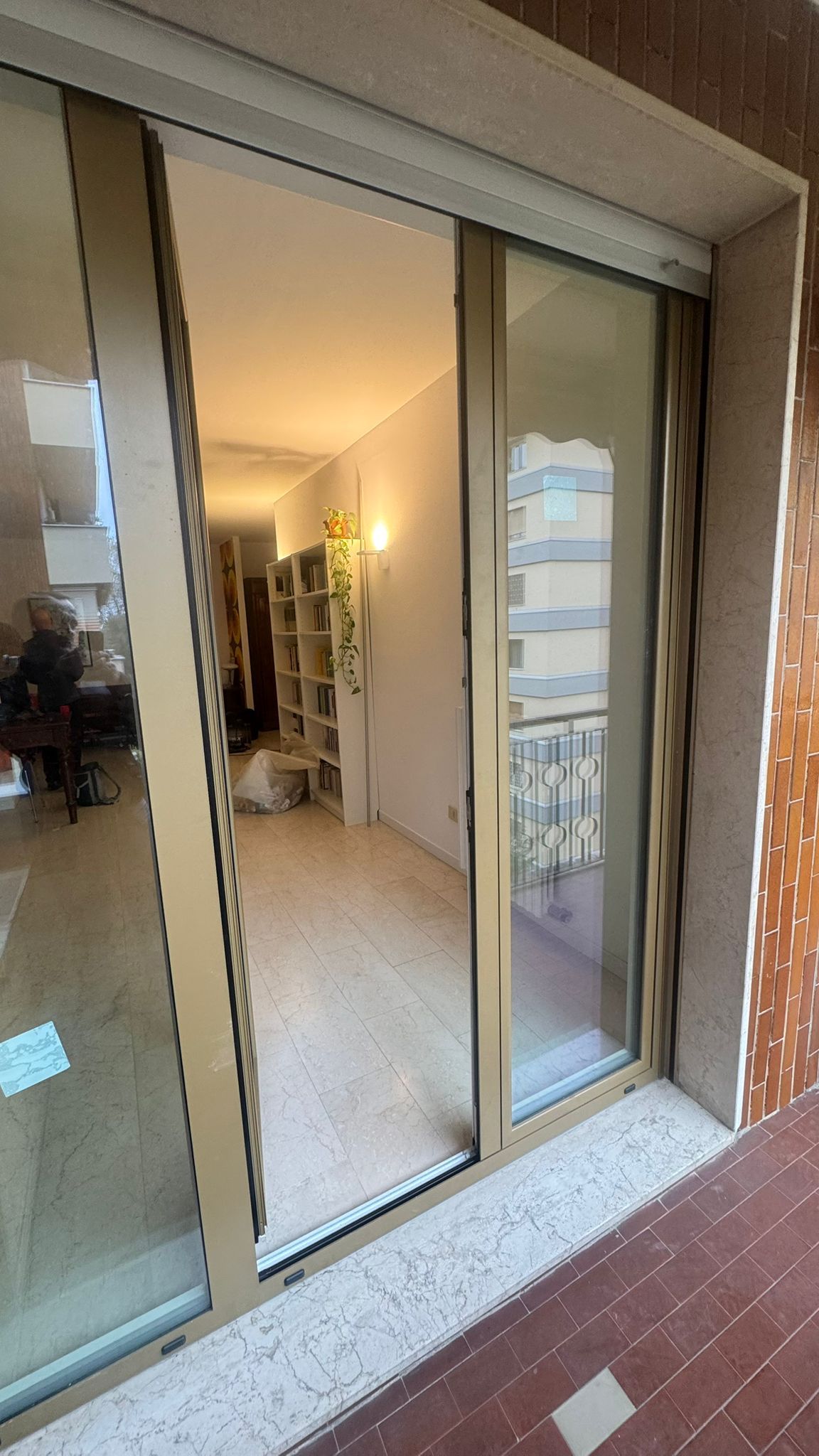 Porta scorrevole in vetro aperta, vista su una stanza interna con pavimento e scaffali chiari. Balcone fuori dalla porta.