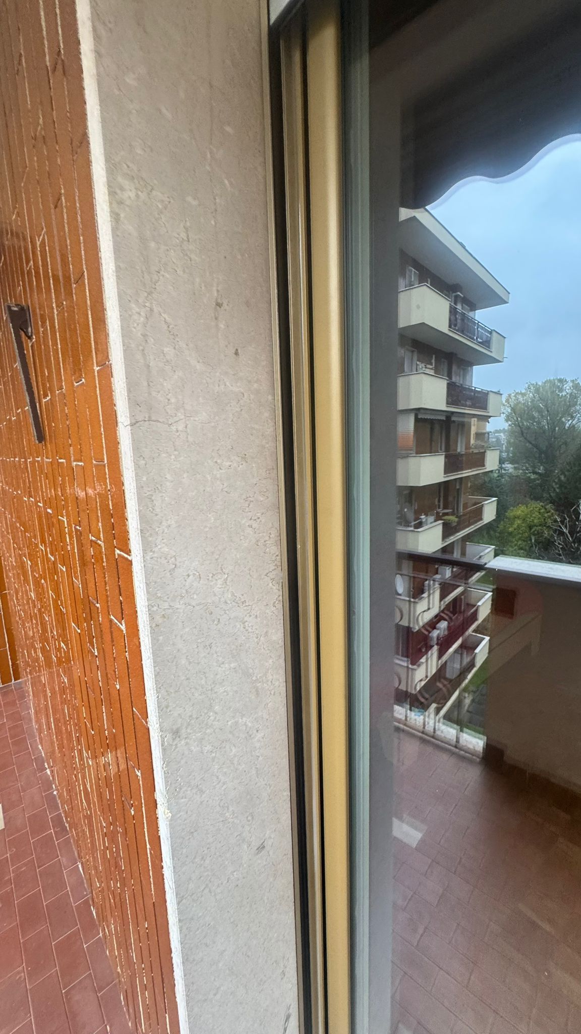 Vista attraverso una finestra, con un balcone, un edificio e un cielo nuvoloso. Toni beige e marroni.