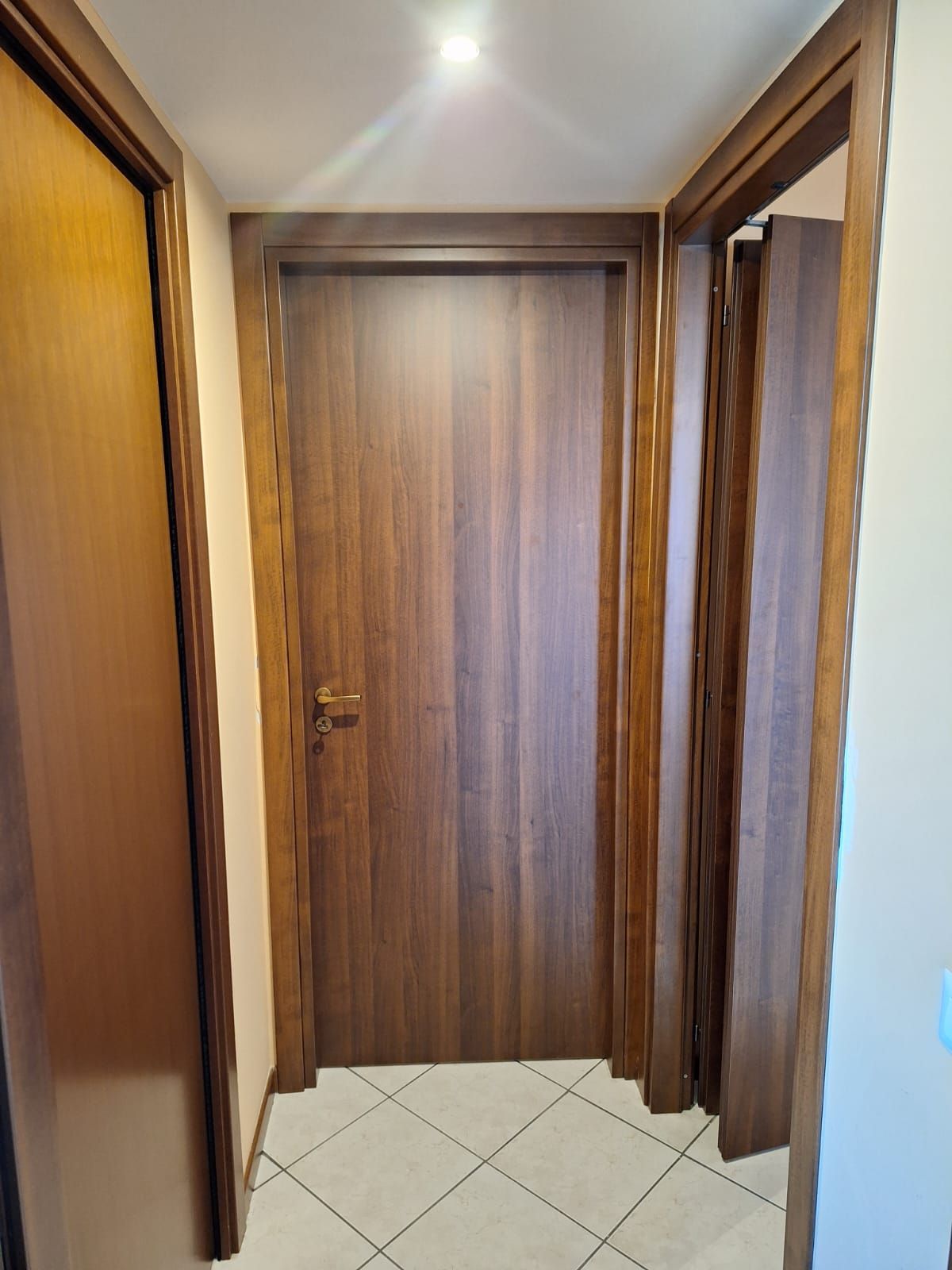 Porte di legno marrone in un corridoio, una chiusa e una aperta. Pareti beige, pavimento piastrellato.