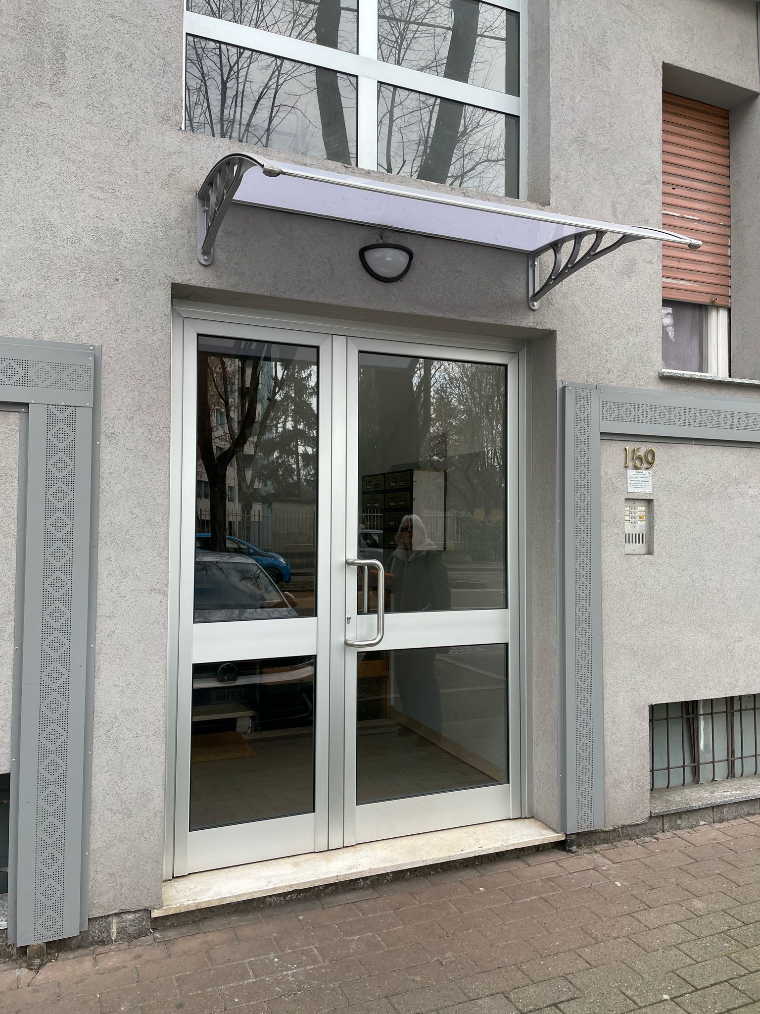 Porte doppie in vetro con telai in alluminio, sotto una piccola pensilina, in un edificio in stucco grigio.