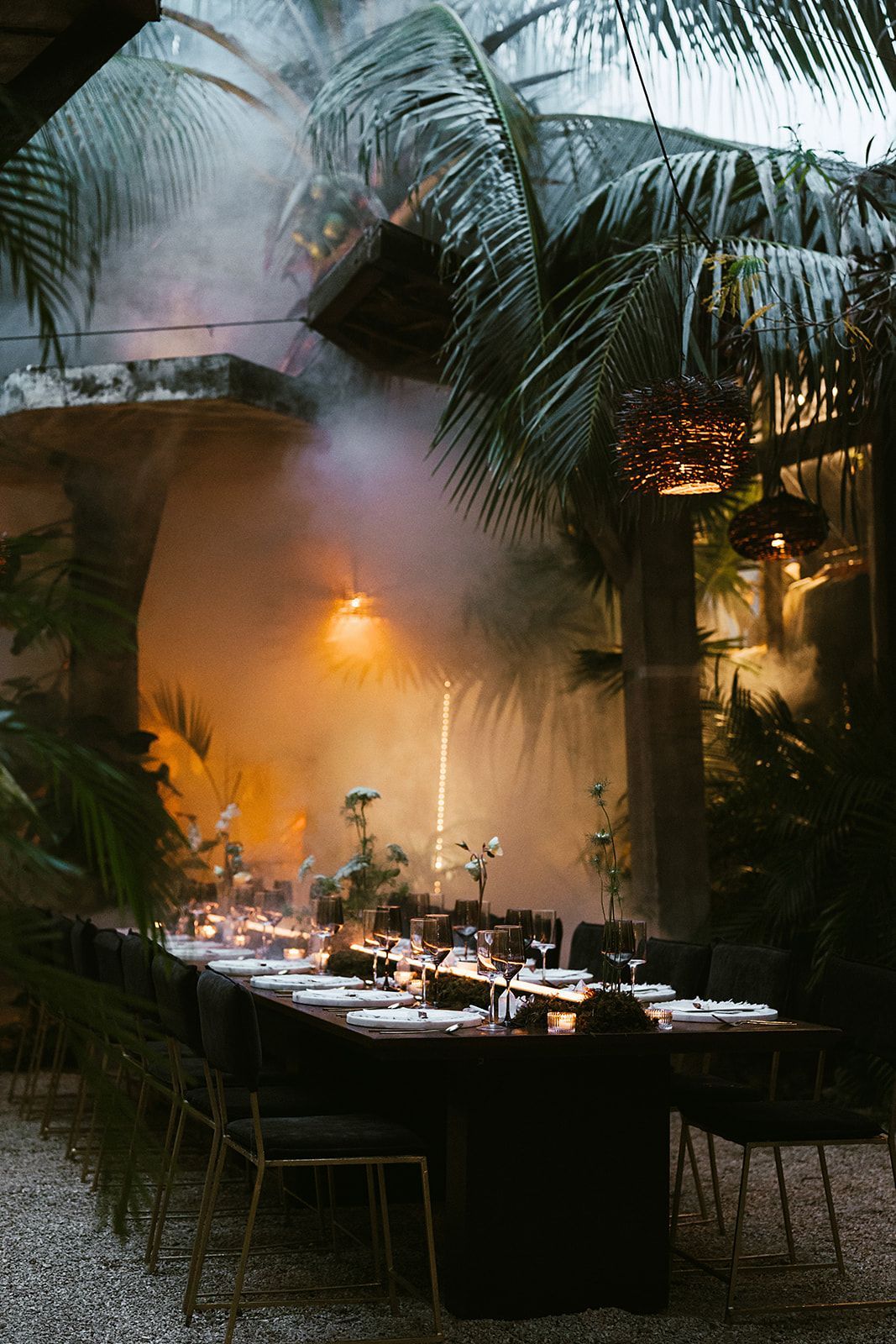 Long table set for a dinner party in an outdoor space, surrounded by greenery and smoke.