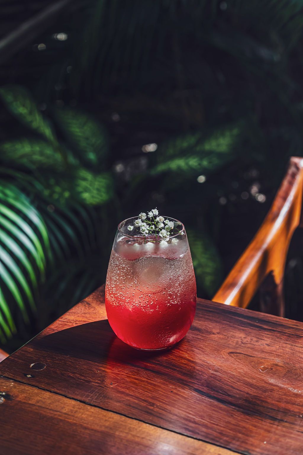 Cocktail in a stemless glass, red base, clear fizz, flower garnish, on a wooden table, green foliage background.