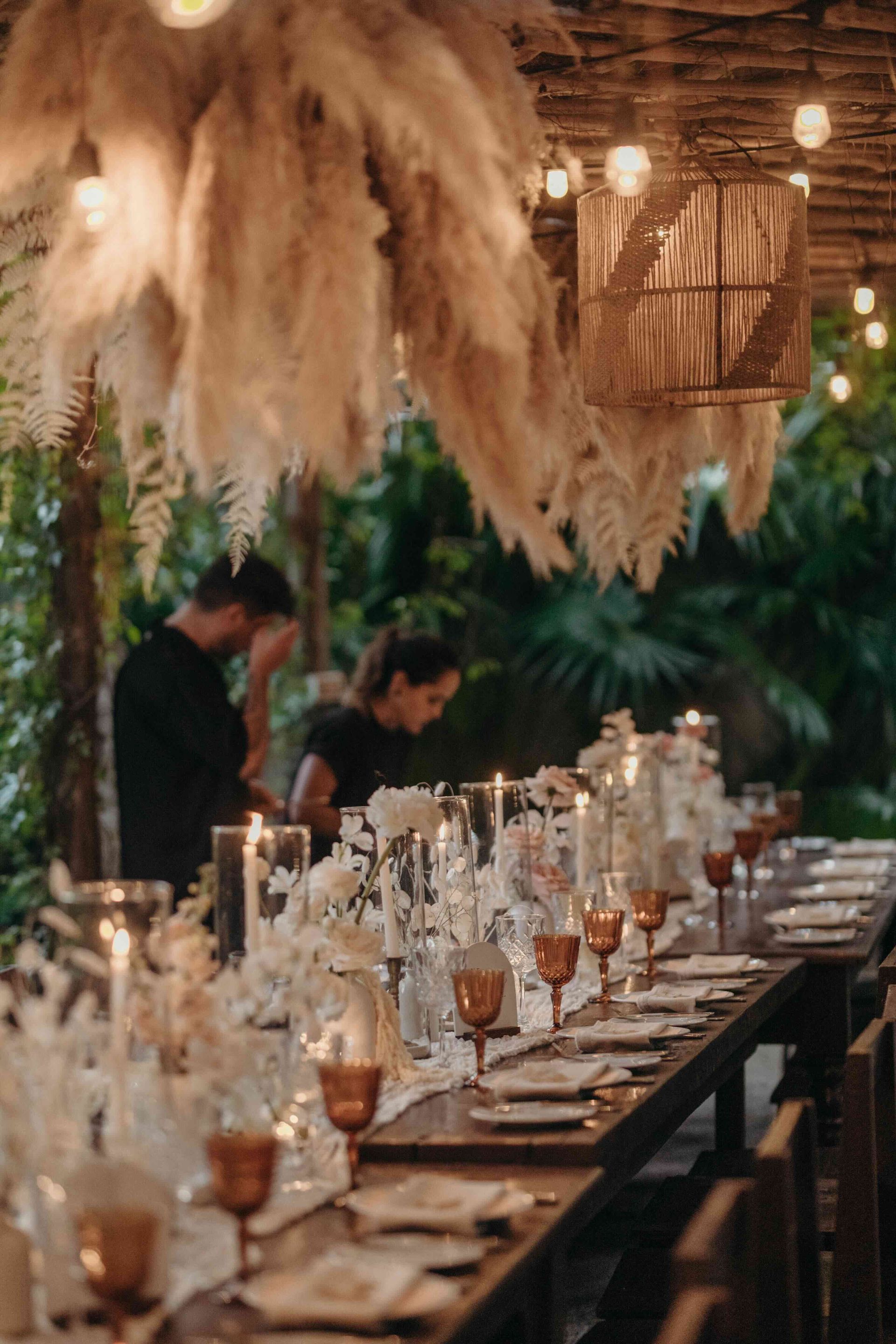 Long dining table set for a wedding with amber glassware, white floral decorations, and pampas grass.