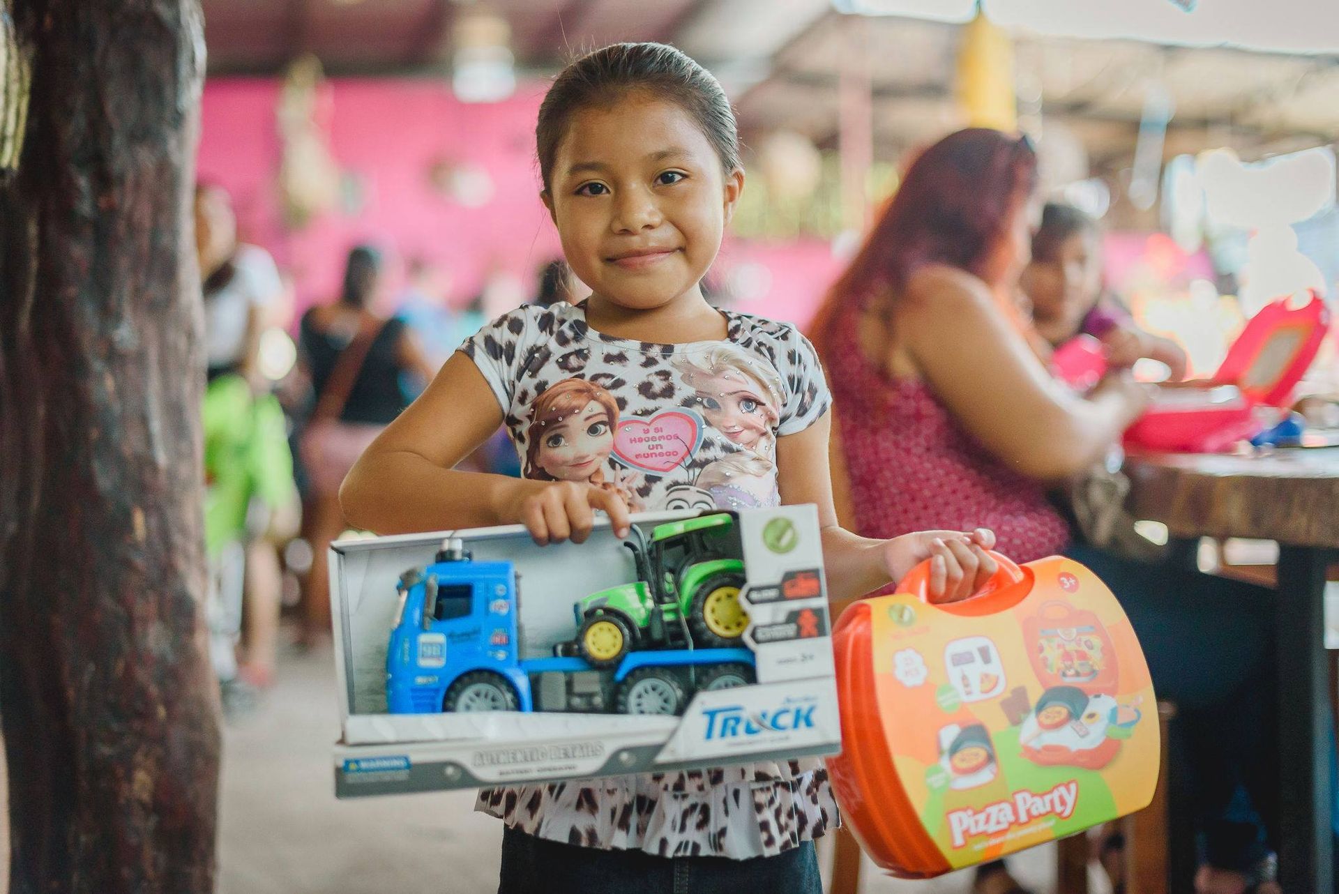 A little girl is holding a toy truck and a lunch box.