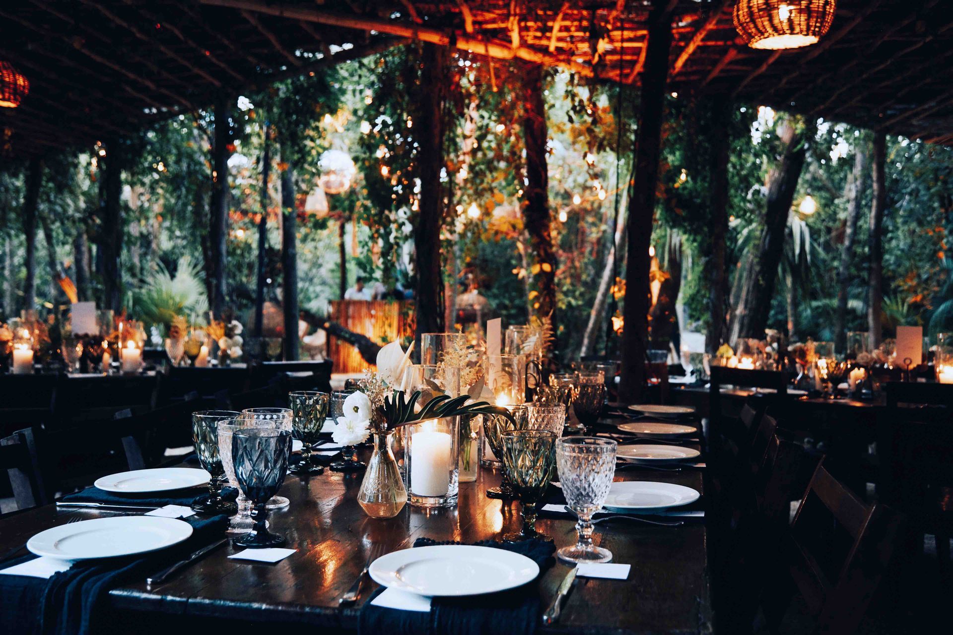 A long table with plates , glasses , and candles in a room with trees in the background.