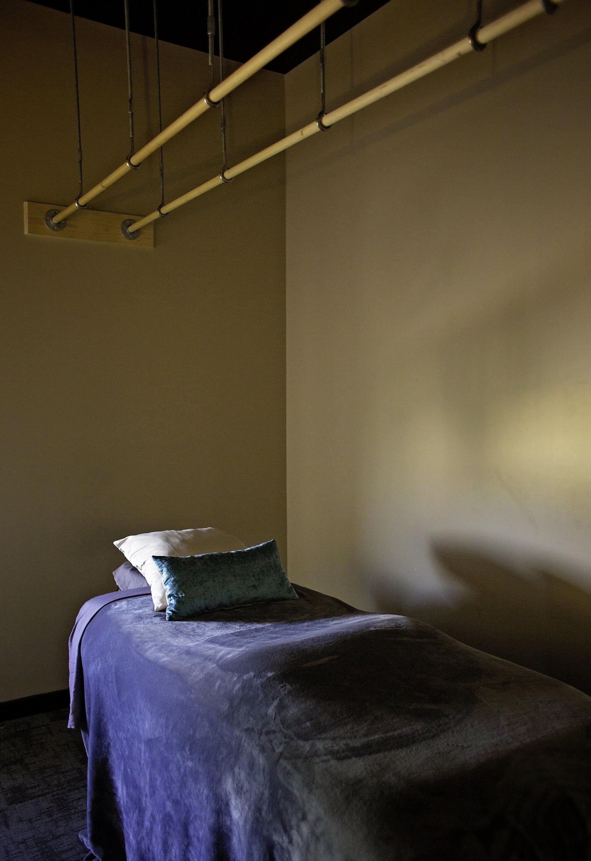 A bed with a purple blanket and pillows in a room with pipes hanging from the ceiling.