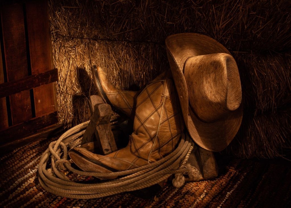 Cowboy hat, boot, and rope rest in a dimly lit barn setting.