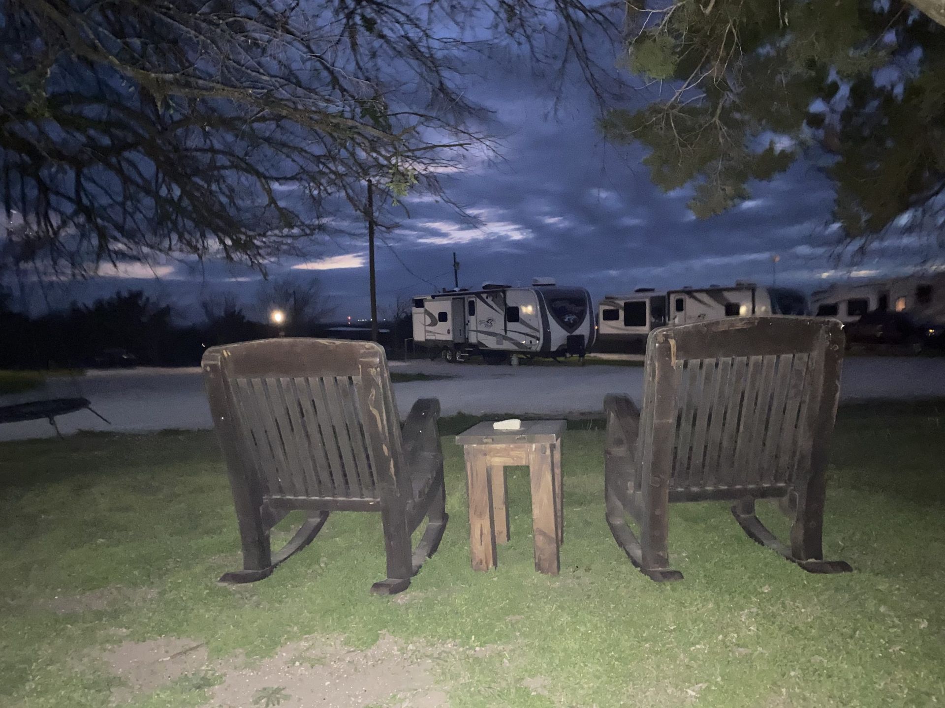 Two wooden rocking chairs face away from the viewer towards RVs in a campground at dusk.