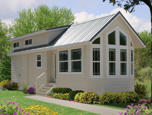 Beige tiny house with white trim, metal roof, and large front windows, set in a yard.