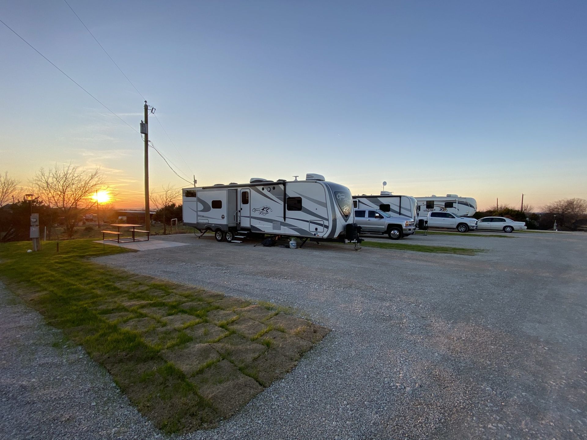 Campground with RVs parked on gravel, sunset in background, clear sky.