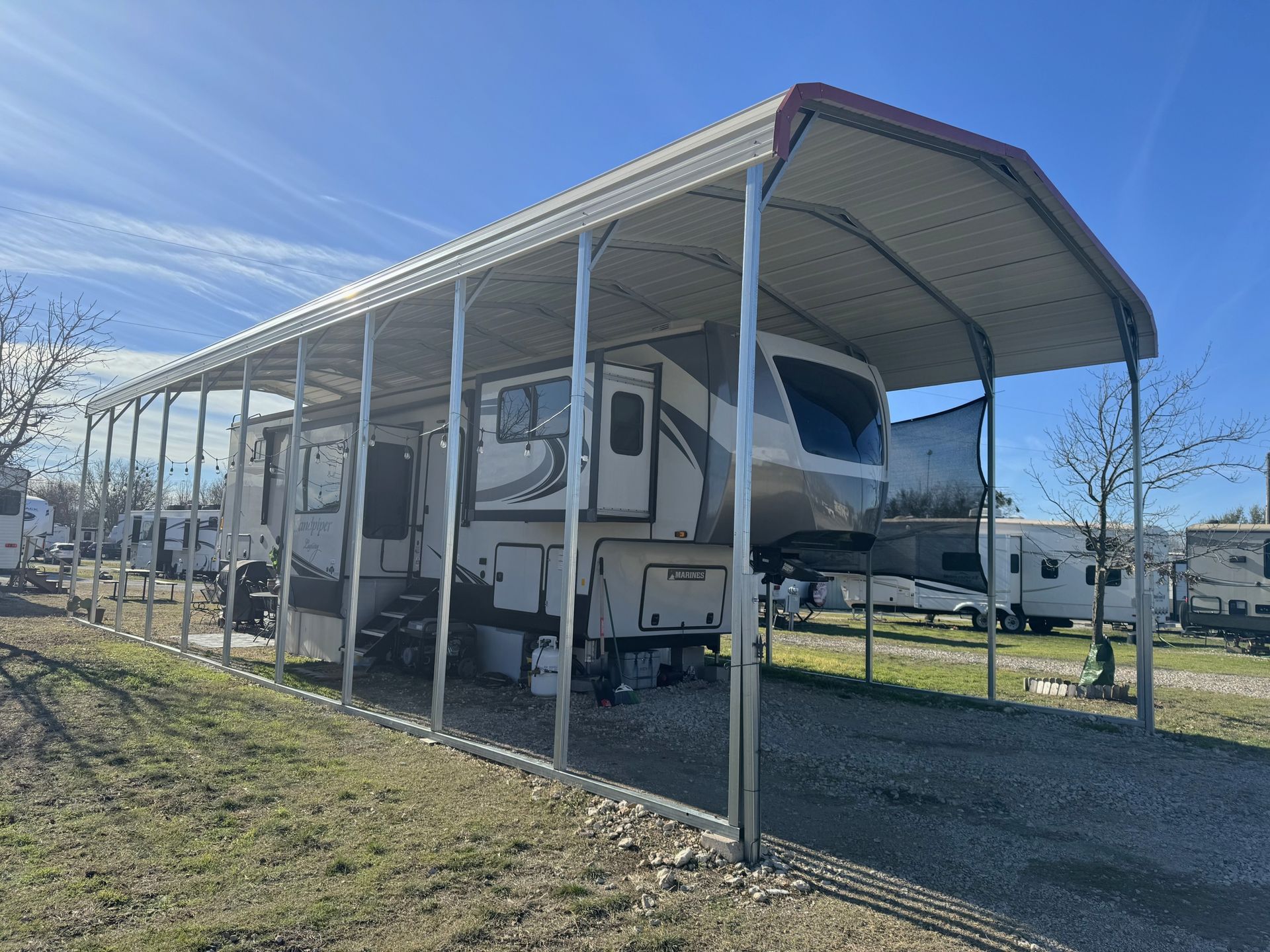 RV parked under a metal carport in a grassy lot; other RVs visible in background on a sunny day.