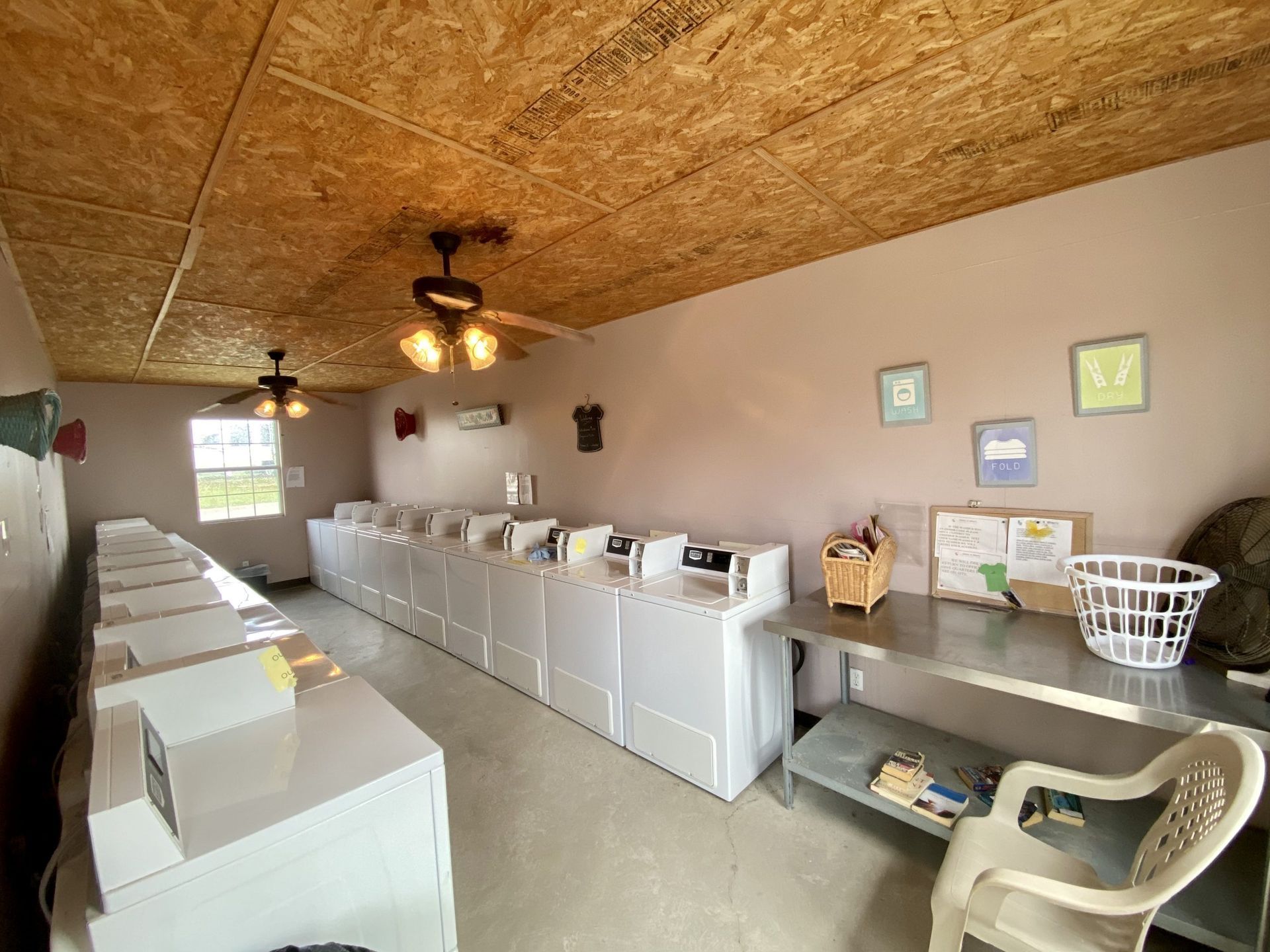 A laundromat with rows of white washing machines and folding tables. Brown ceiling with two fans.