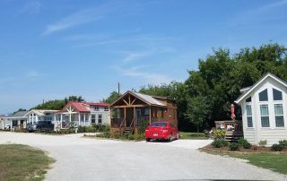 Tiny houses on a gravel road, with a red car parked near one. Blue sky and green trees.