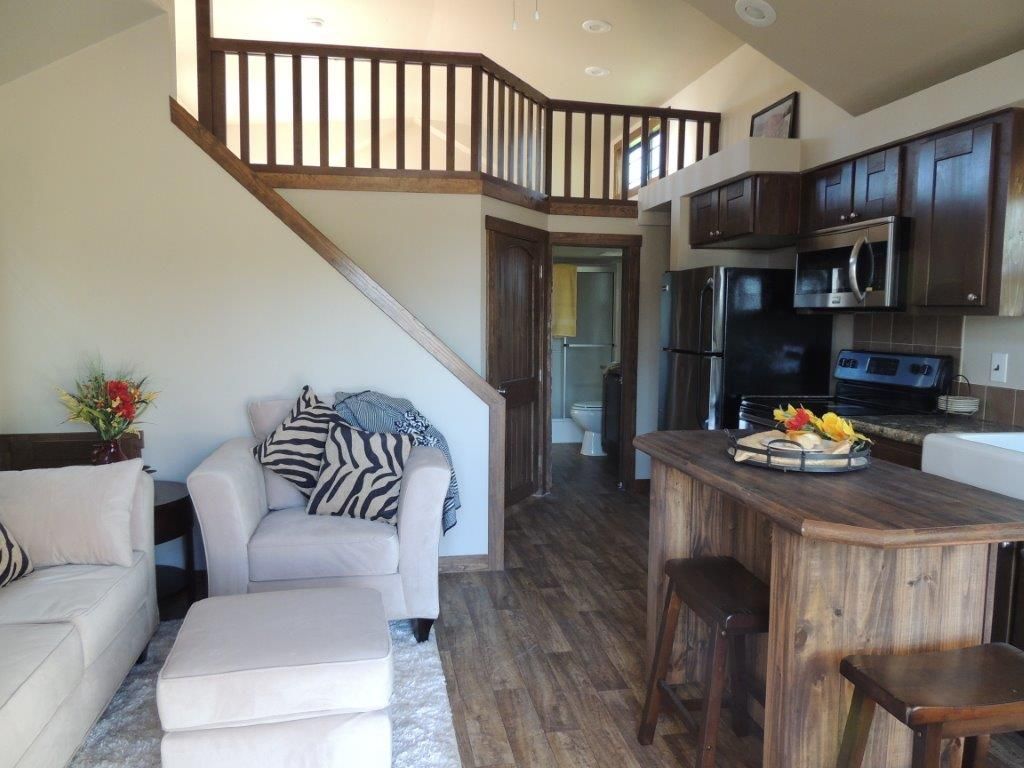 Interior of a tiny home with a loft, kitchen, living area, and bathroom visible. Brown cabinetry and wood flooring.