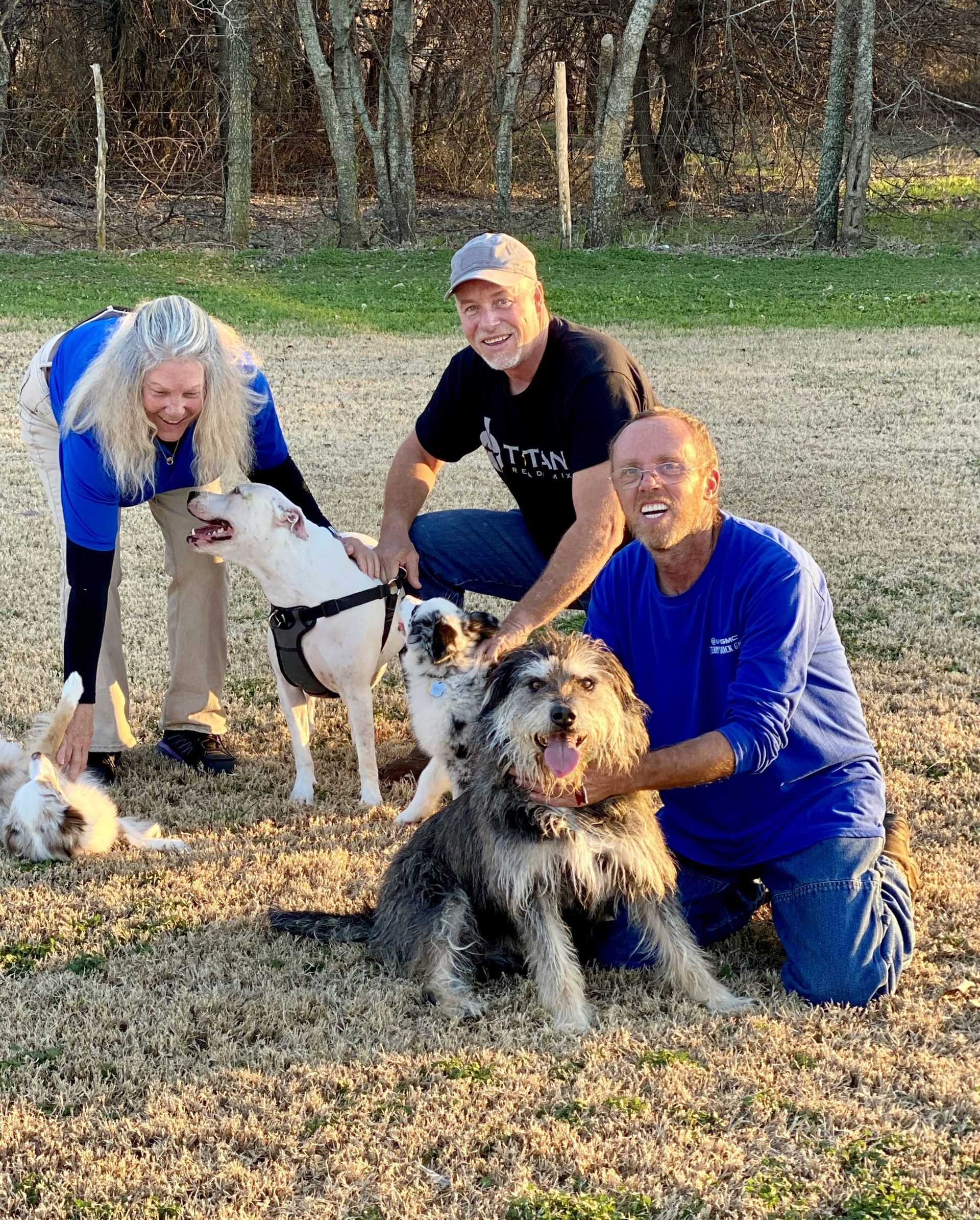Three people kneeling with four dogs in a grassy area, smiling.
