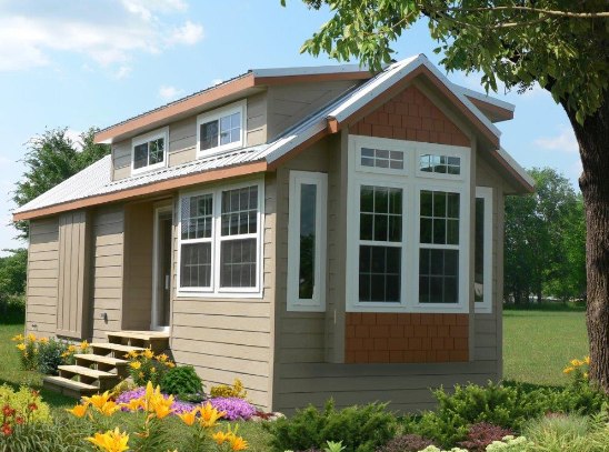 Tan tiny house with white trim, bay window, and gabled roof, nestled among flowers and trees.