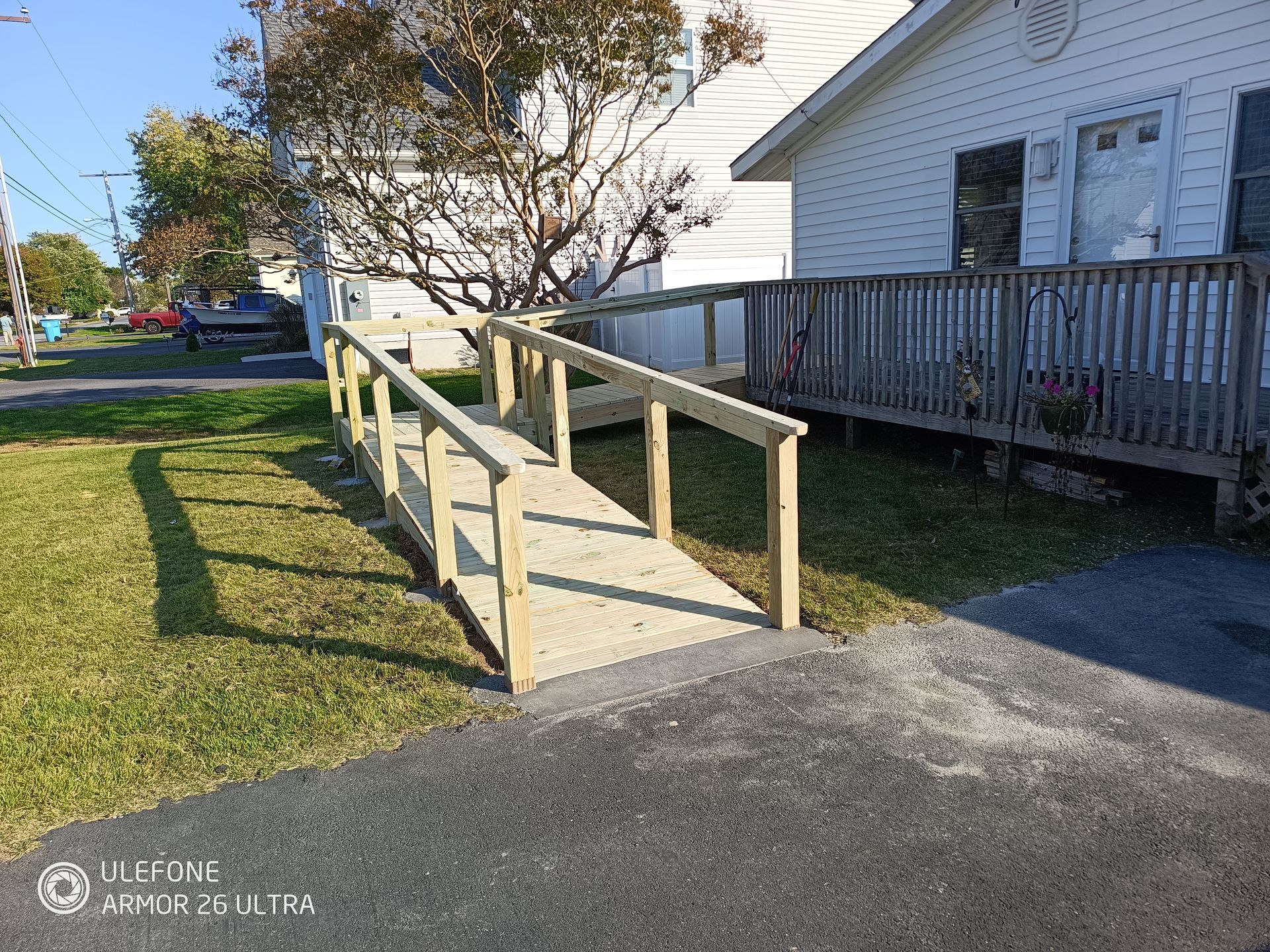 Wooden ramp leading to a white building with a deck; green grass and a paved driveway are in the foreground.