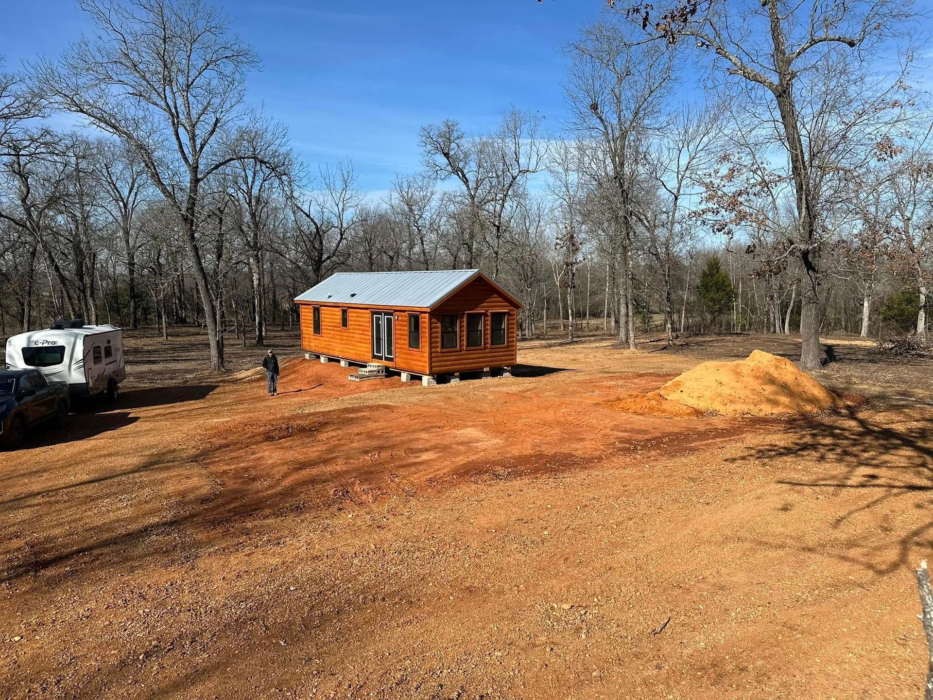 Small wooden cabin on a cleared, reddish dirt lot surrounded by leafless trees.