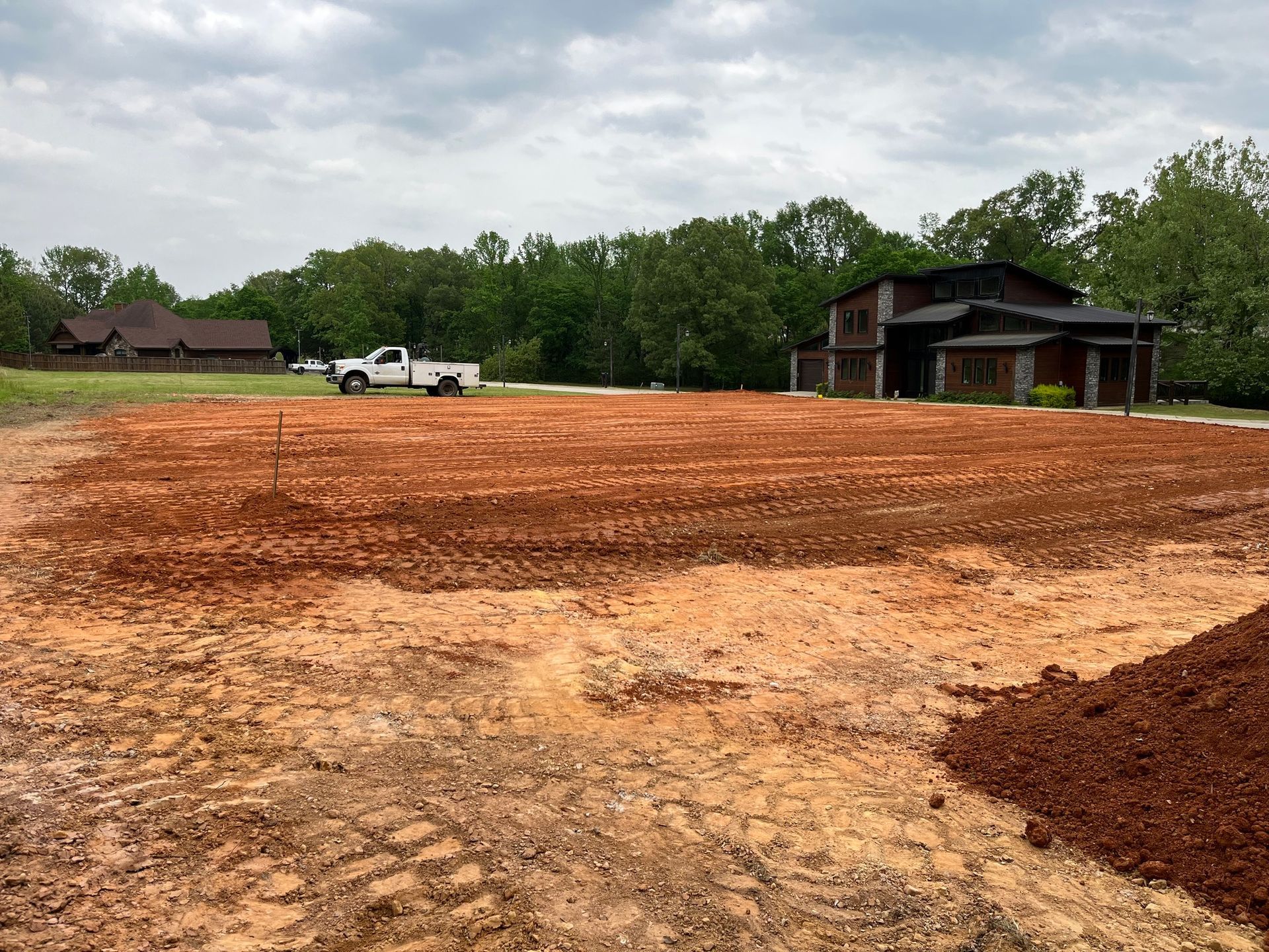 A cleared red-earth lot with a truck, ready for construction, with a house in the background under a cloudy sky.