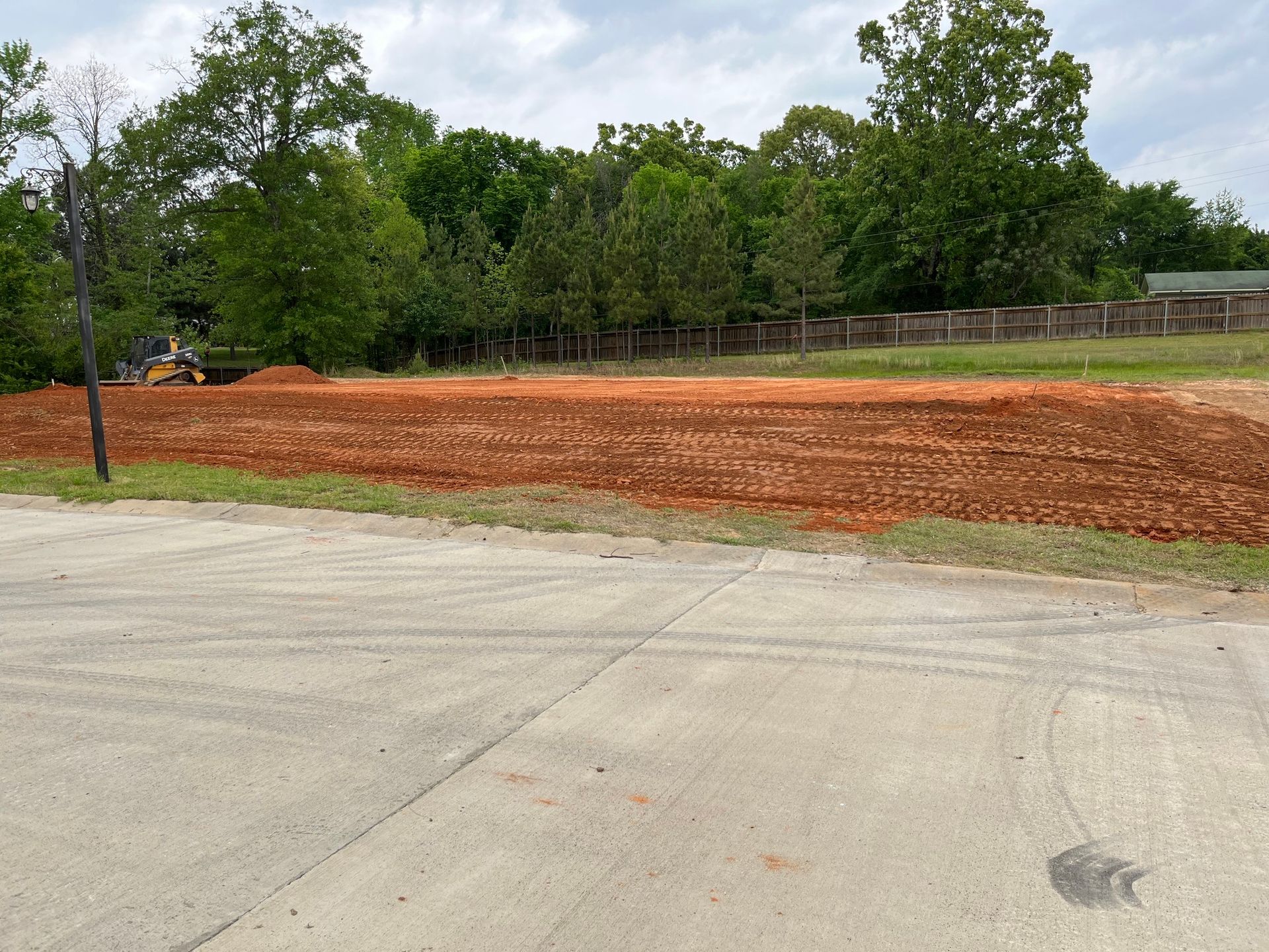 A leveled, reddish-brown dirt lot with trees in the background, seen from a gray asphalt road on an overcast day.