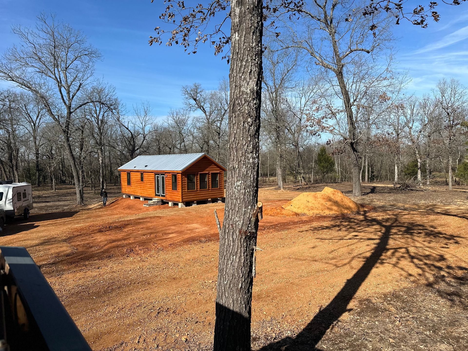 Wooden cabin in a clearing with a dirt ground and trees. Construction materials and a truck are nearby.